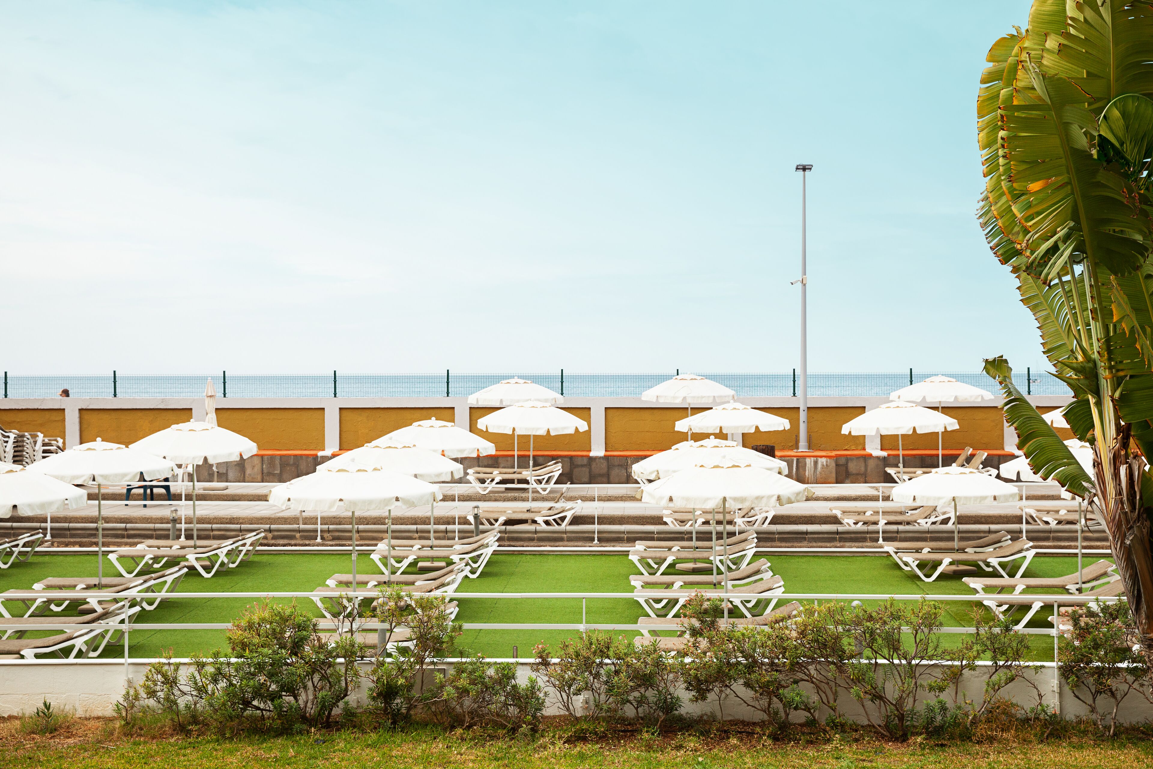 A poolside area with white lounge chairs and umbrellas, a green artificial turf, and a view of the sea.