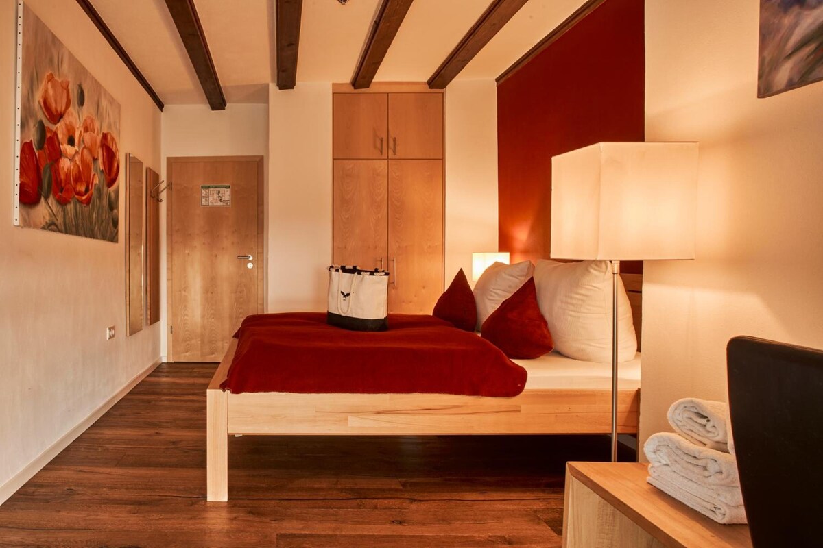 Une chambre d’hôtel avec un lit en bois, un plaid rouge, une lampe de chevet et un tableau représentant des fleurs accroché au mur.