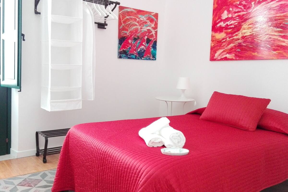 Ein Zimmer mit einem Bett, einem weißen Handtuch, einer roten Decke, einem kleinen Tisch mit einer Lampe und einem Gemälde an der Wand.