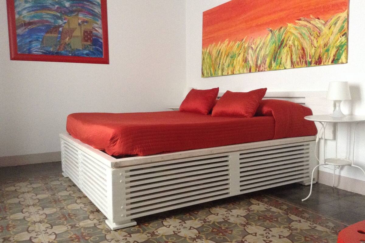 Ein Bett mit roter Bettdecke und Kissen, ein gemusterter Boden, eine weiße Heizung und zwei Bilder an der Wand.