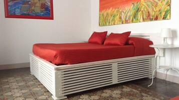 Un letto con copriletto e cuscini rossi, un pavimento a motivi, un termosifone bianco e due quadri appesi al muro.