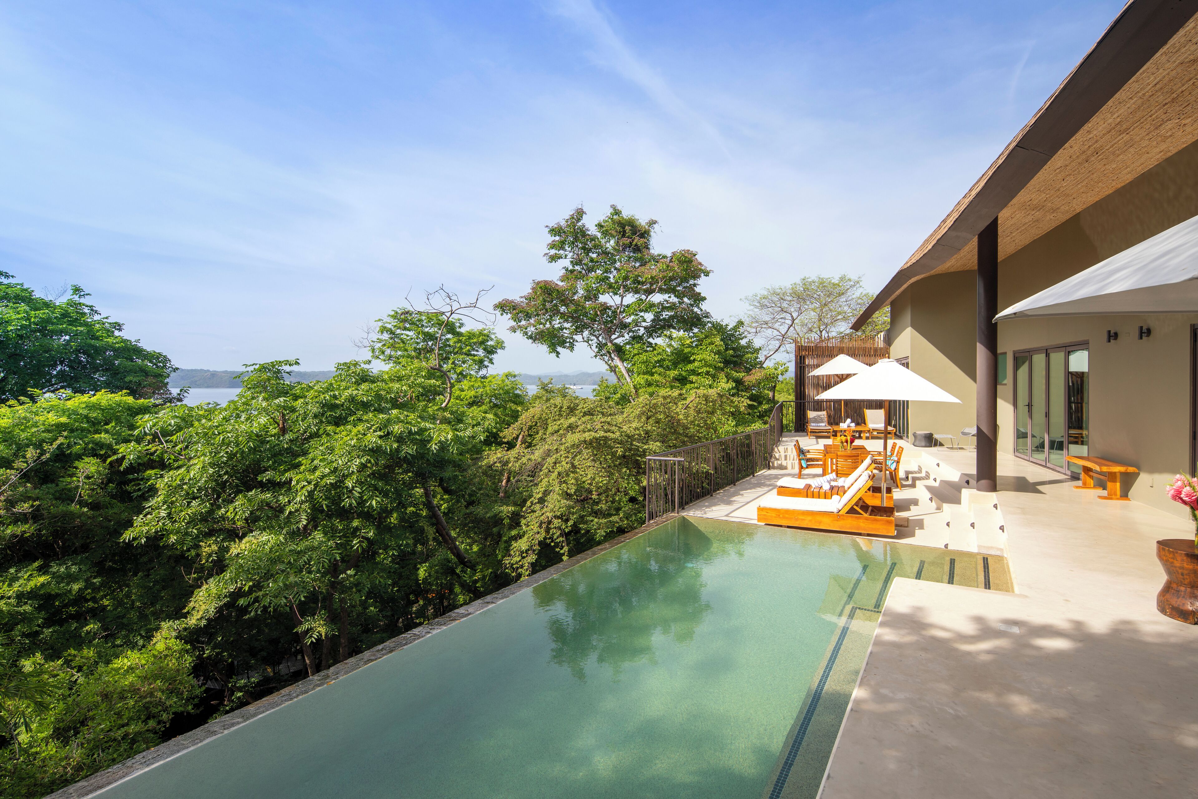 A poolside area with a view of the sea, surrounded by lush greenery and modern outdoor furniture.
