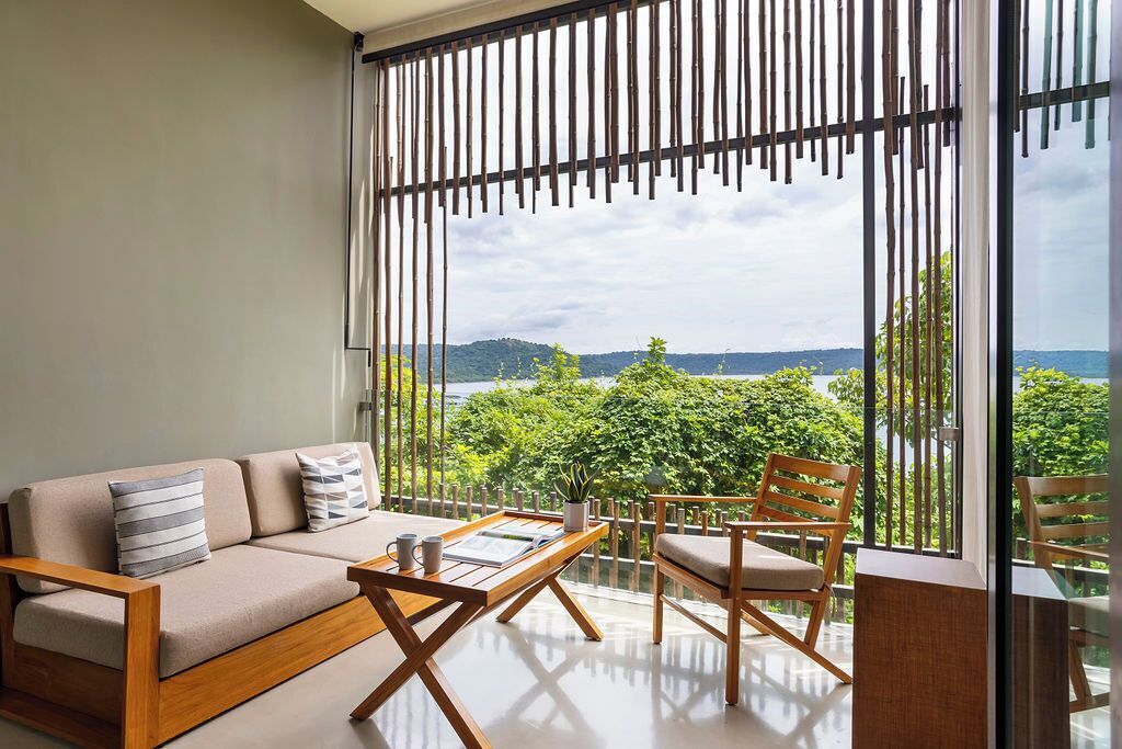 A modern living room with a sofa, coffee table, and chairs, large windows with a view of greenery and hills.