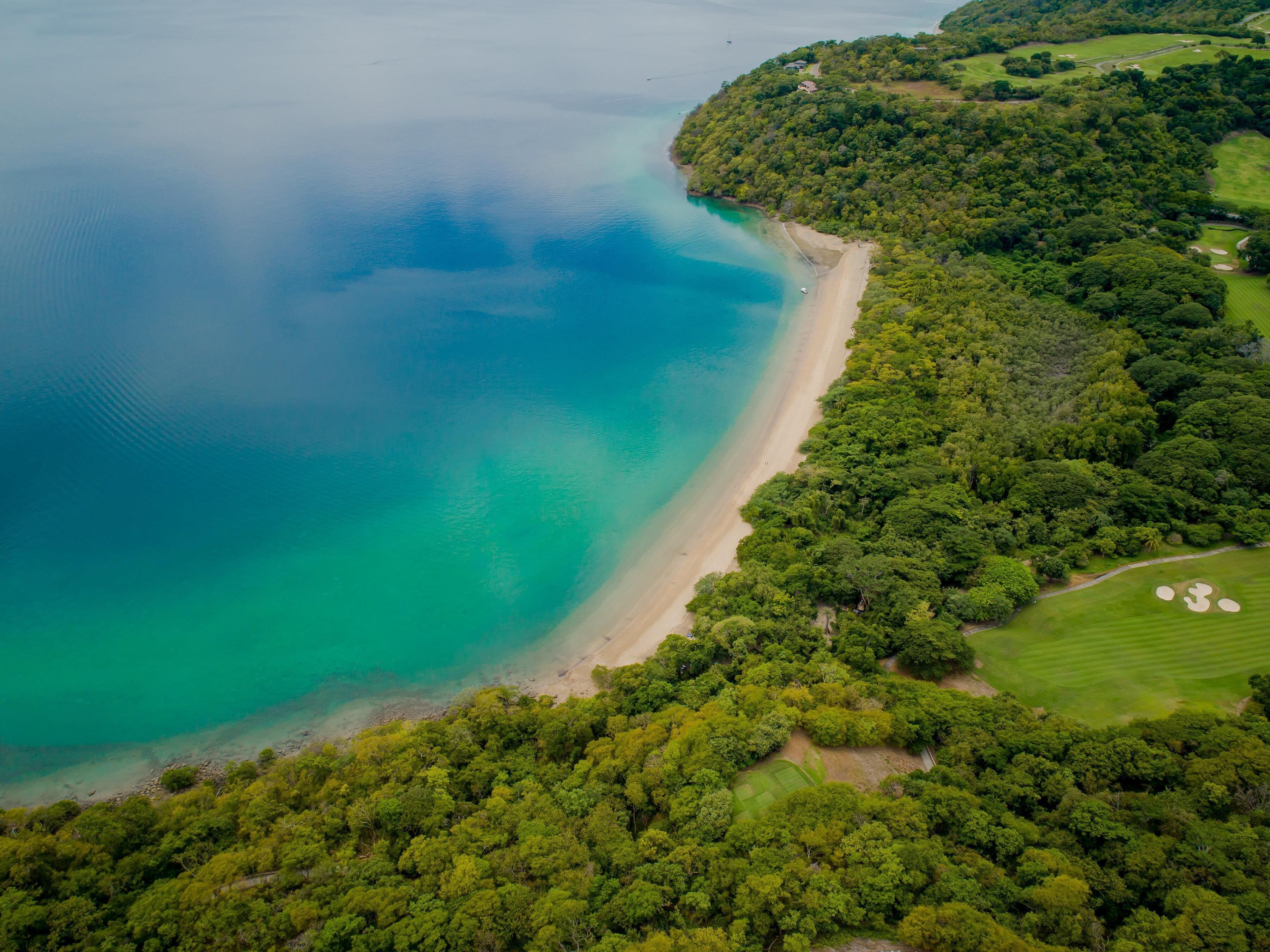An aerial view of a coastal area with a beach, dense greenery, and golf course.