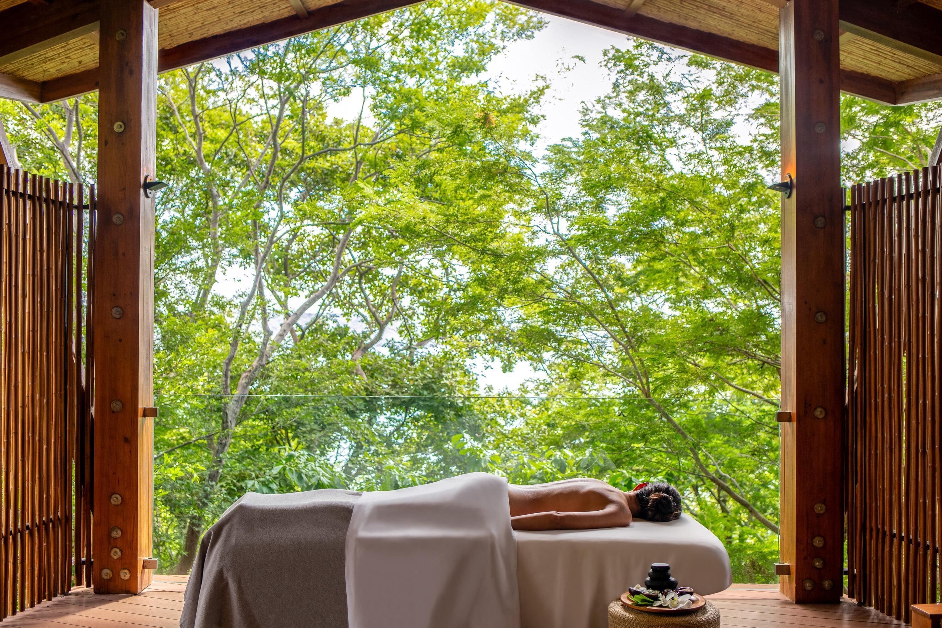 A wooden deck with a person lying on a massage table, surrounded by vertical wooden slats and a view of green trees.