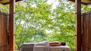 A wooden deck with a person lying on a massage table, surrounded by vertical wooden slats and a view of green trees.