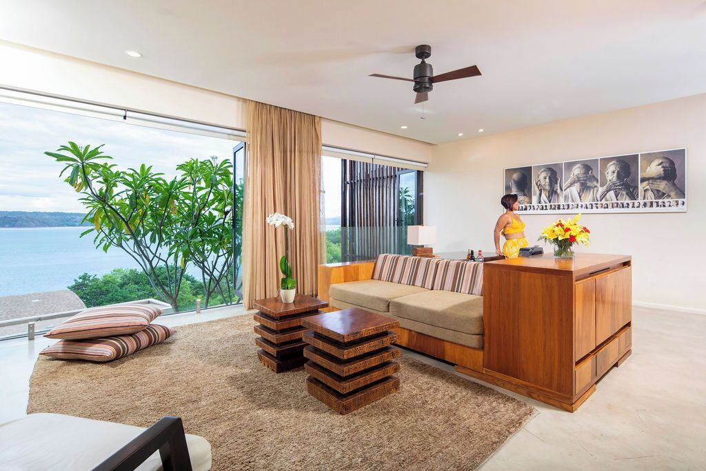 Ein modernes Wohnzimmer mit einer Couch, einem Couchtisch und einer Holzkabine. Ein großes Fenster bietet Blick auf das Meer und viel Grün.