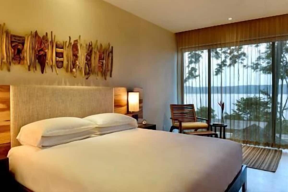 Ein Hotelzimmer mit einem großen Bett, einem Nachttisch mit Lampe, einem Sessel und Blick ins Freie.