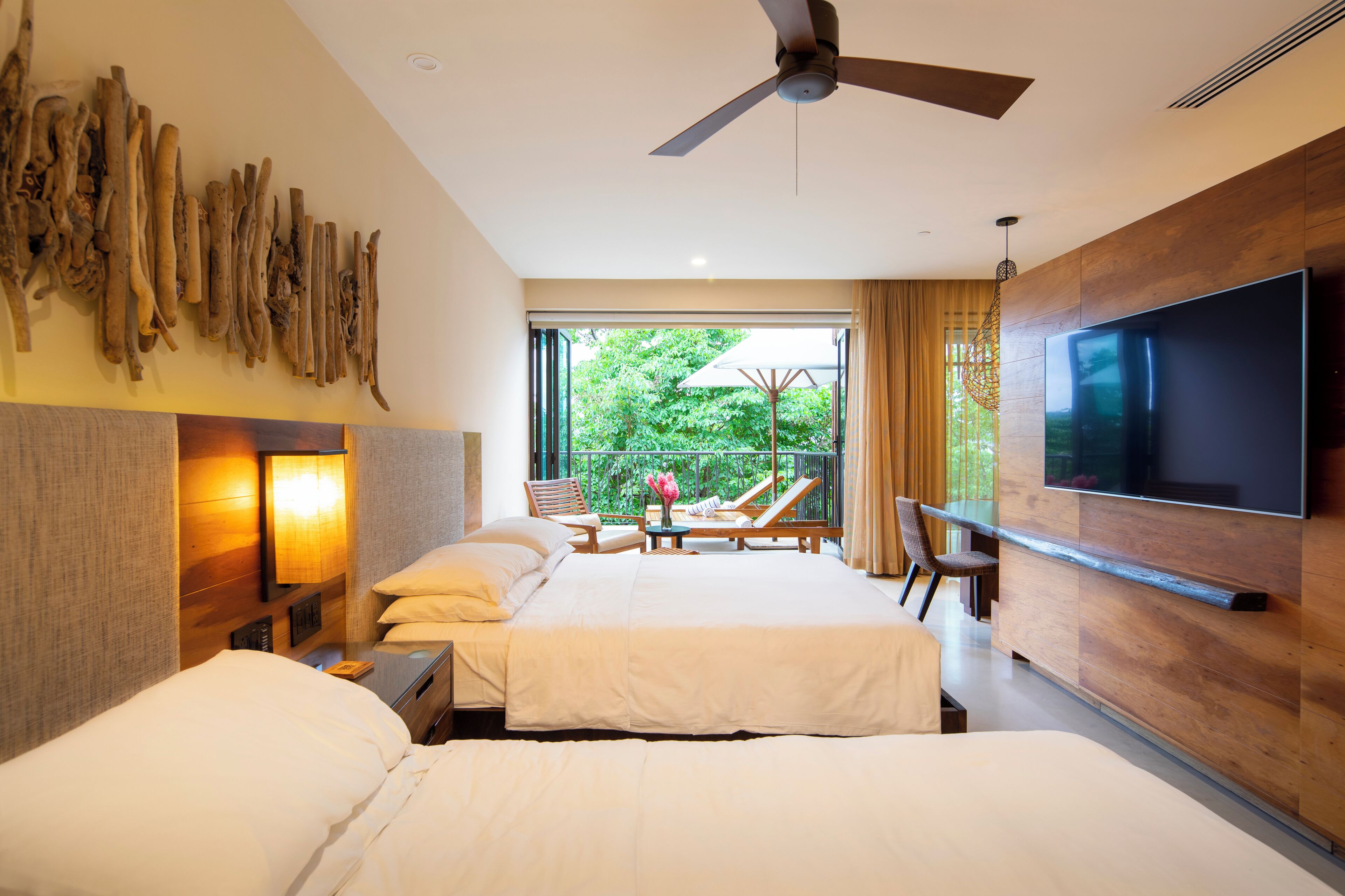A modern hotel room with two beds, a flat-screen TV, a ceiling fan, and a balcony with a view of greenery.