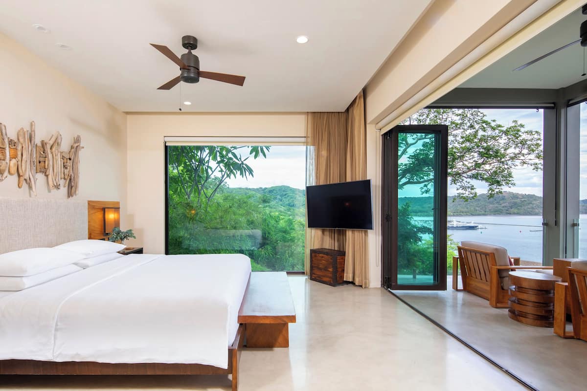 Ein modernes Hotelzimmer mit einem großen Bett, einem Deckenventilator, einem Fernseher und Blick auf einen See und Bäume.