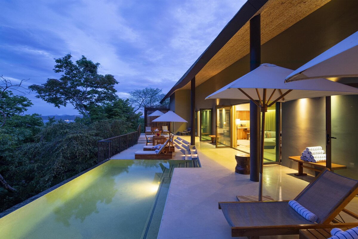 Un área de piscina moderna al aire libre con sillones de descanso y sombrillas, una habitación con paredes de vidrio y cama, y vistas del paisaje circundante.