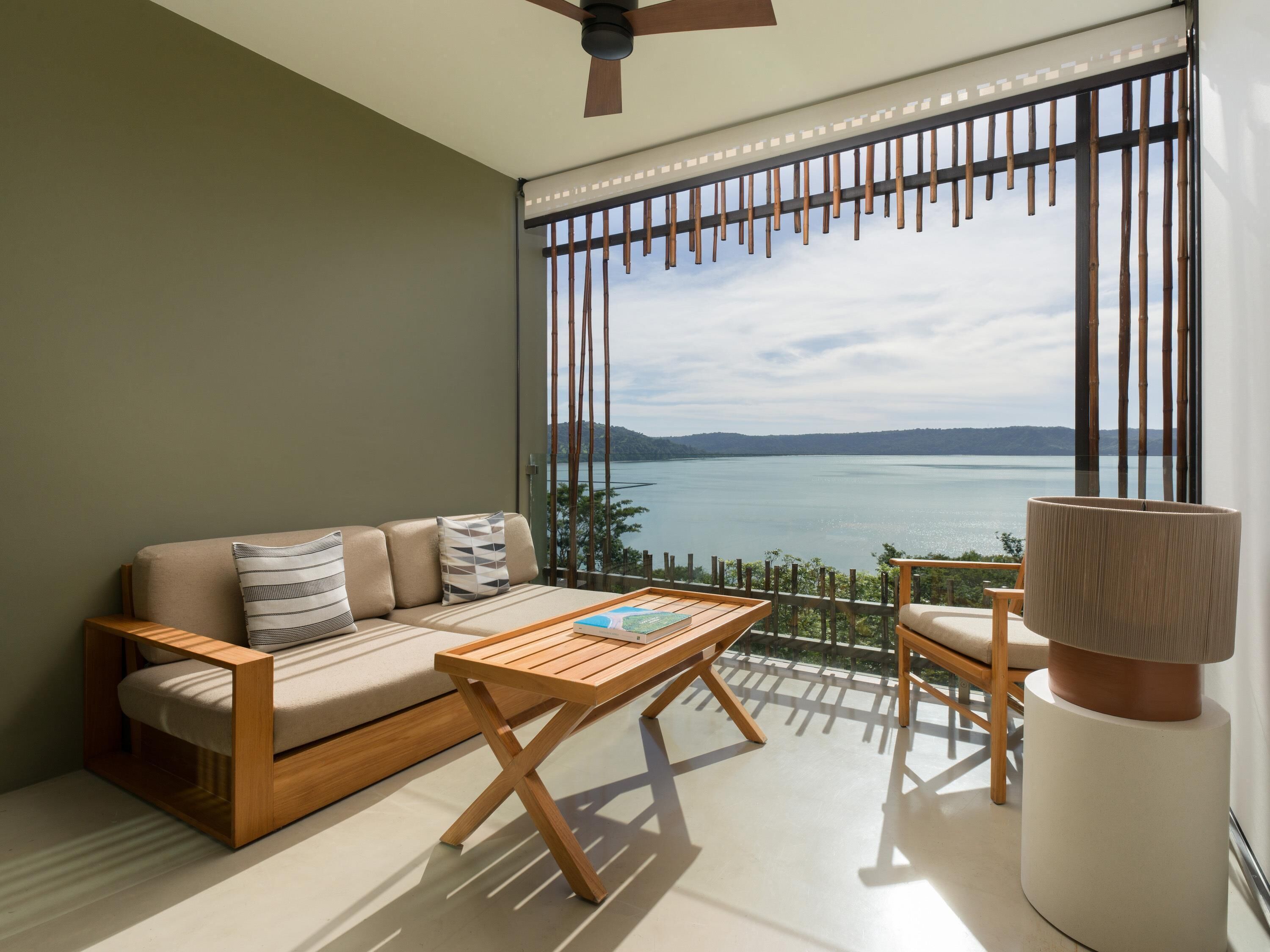 Un balcón con sofá, mesa de madera y una silla, con vistas a un cuerpo de agua y colinas.