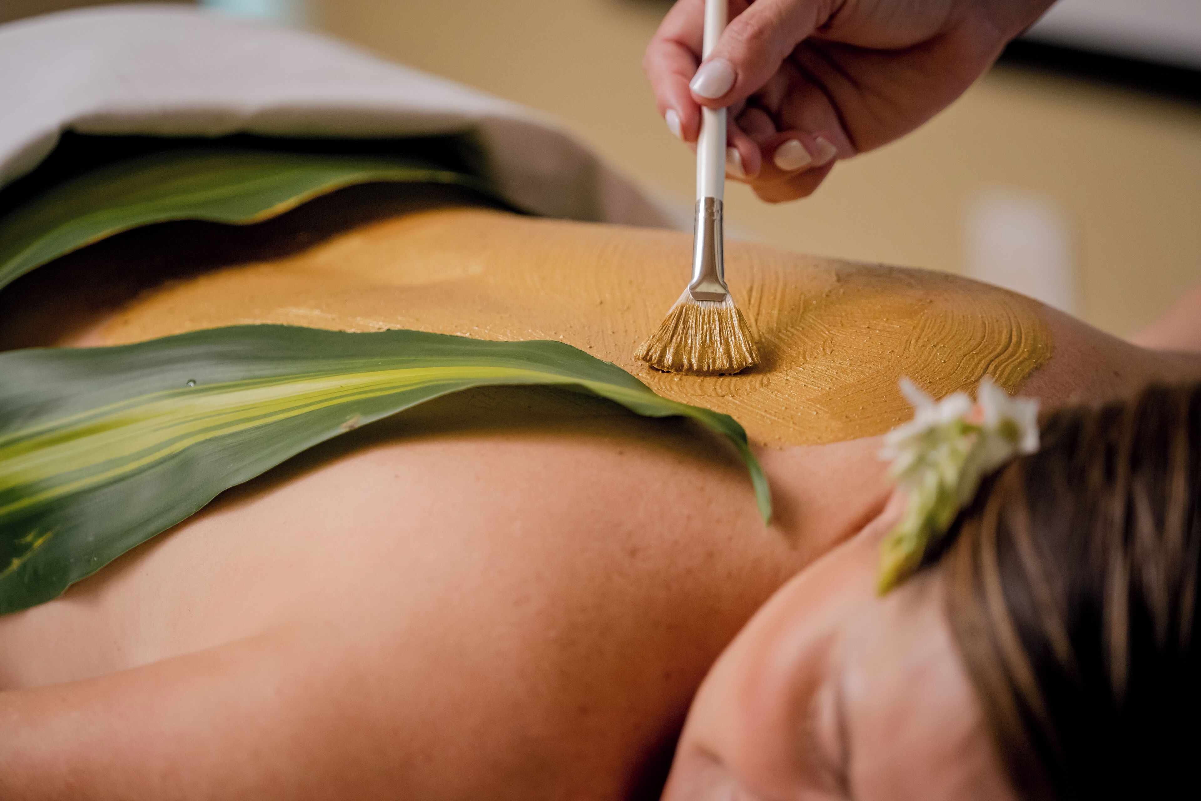 Une personne reçoit un massage du dos à l’aide d’une brosse, porte une feuille verte et a une fleur dans les cheveux.