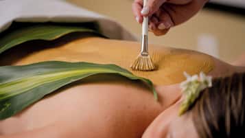 Une personne reçoit un massage du dos à l’aide d’une brosse, porte une feuille verte et a une fleur dans les cheveux.