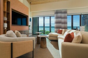 A modern living room with a large TV, comfortable seating, a small table, and a view of the sea.
