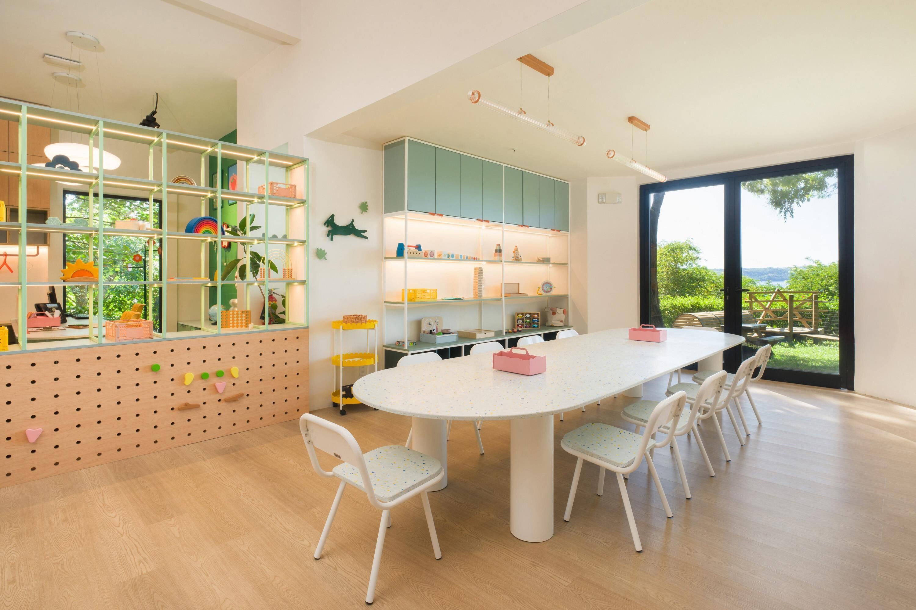 A modern dining area with a round table and white chairs, a children's play area with colorful storage units, and a large window offering a view of greenery.