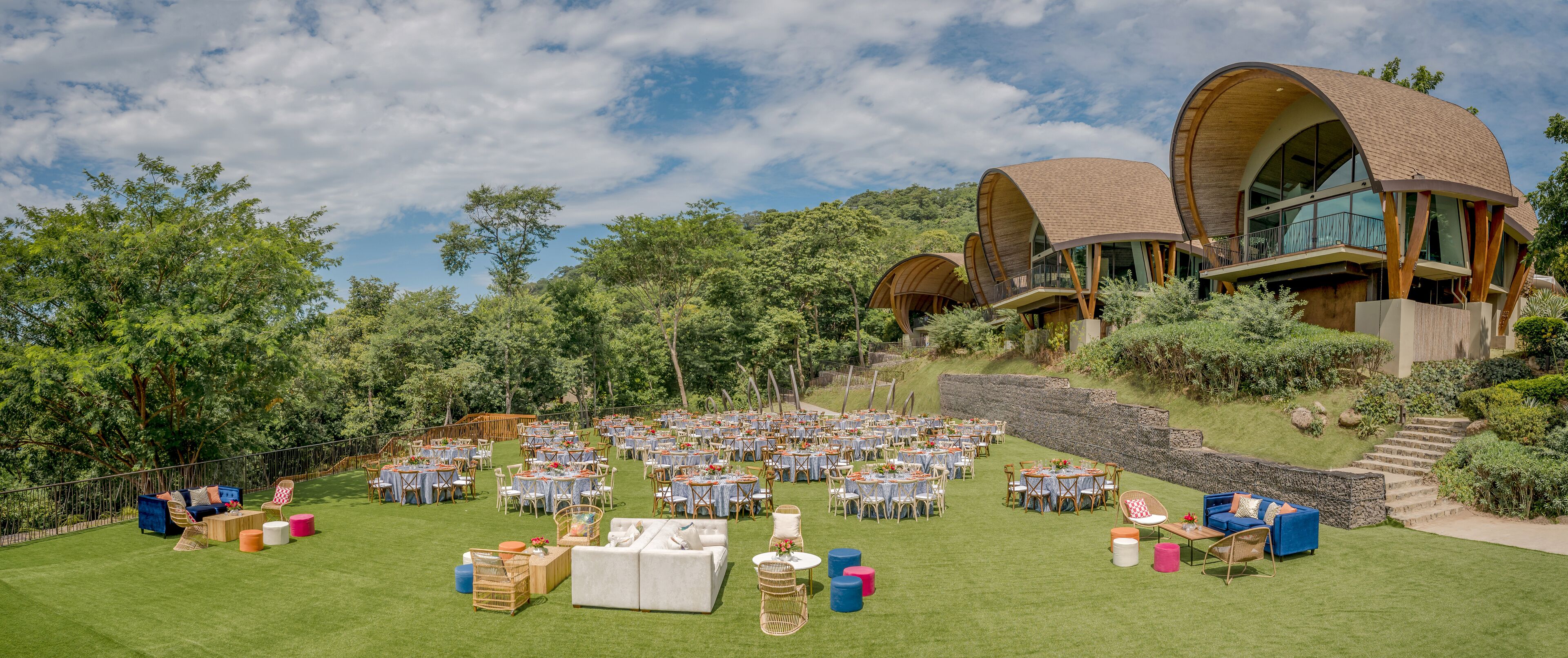 An outdoor event space with tables and chairs, surrounded by lush greenery and a stone wall.
