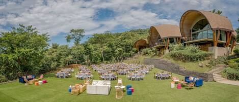 An outdoor event space with tables and chairs, surrounded by lush greenery and a stone wall.