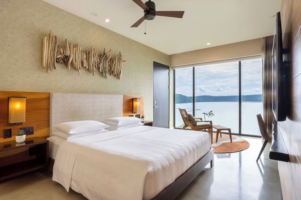 A modern hotel room with a large bed, a ceiling fan, a wall-mounted light, a view of the sea, and a seating area with a table and chairs.