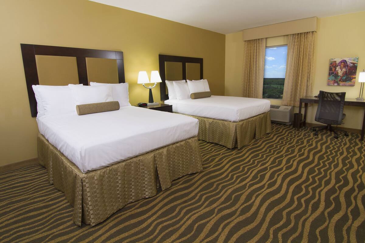 A hotel room with two beds, a desk, a chair, a window with curtains, and a painting on the wall.