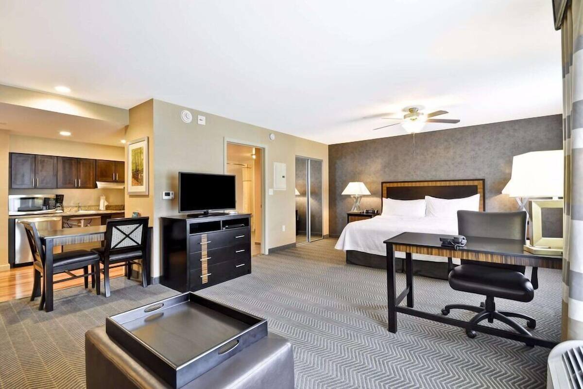 Une chambre d’hôtel équipée d’un lit, d’un bureau, d’une chaise, d’une télévision, d’une kitchenette et d’un ventilateur de plafond.