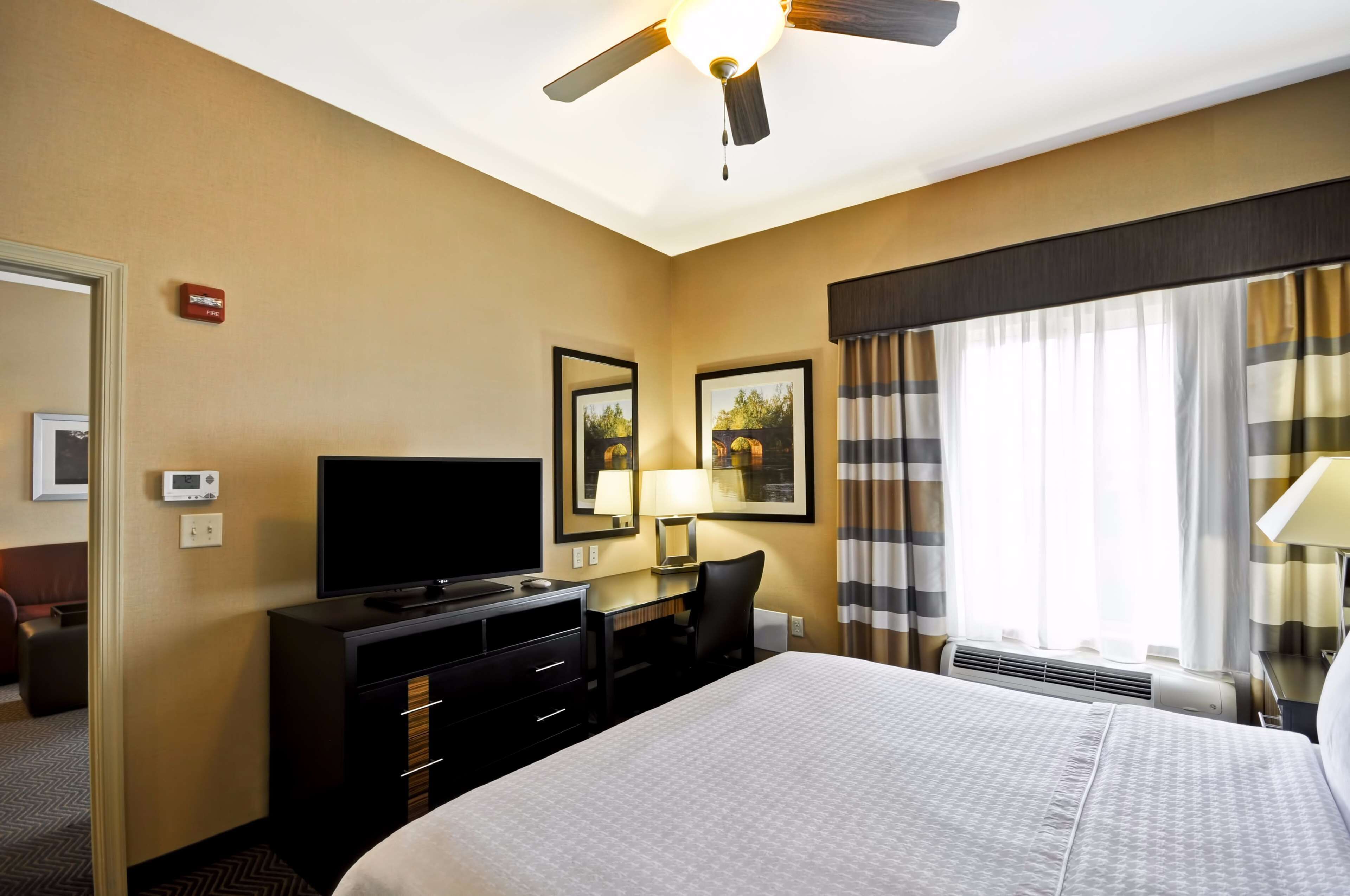 Quarto de hotel com cama, escrivaninha com computador, televisão, ventoinha no teto e janela com cortinas.