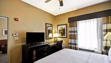 Quarto de hotel com cama, escrivaninha com computador, televisão, ventoinha no teto e janela com cortinas.