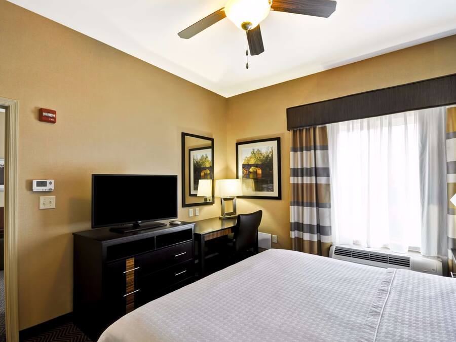 A hotel room with a bed, a desk with a computer, a TV, a ceiling fan, and a window with curtains.
