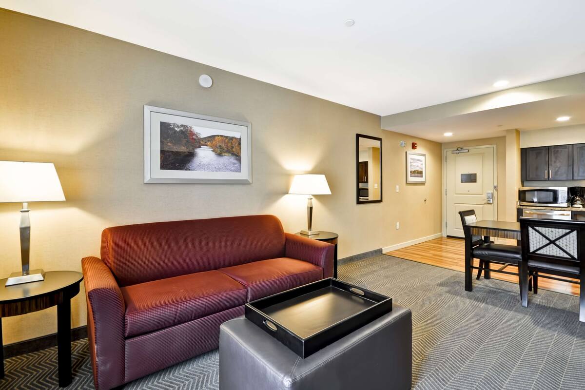 Une chambre d’hôtel avec un canapé rouge, une petite table avec une lampe, une table d’appoint, une table à manger avec des chaises, un coin cuisine et un tableau encadré au mur.