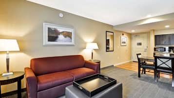 A hotel room with a red sofa, a small table with a lamp, a tray table, a dining table with chairs, a kitchen area, and a framed picture on the wall.