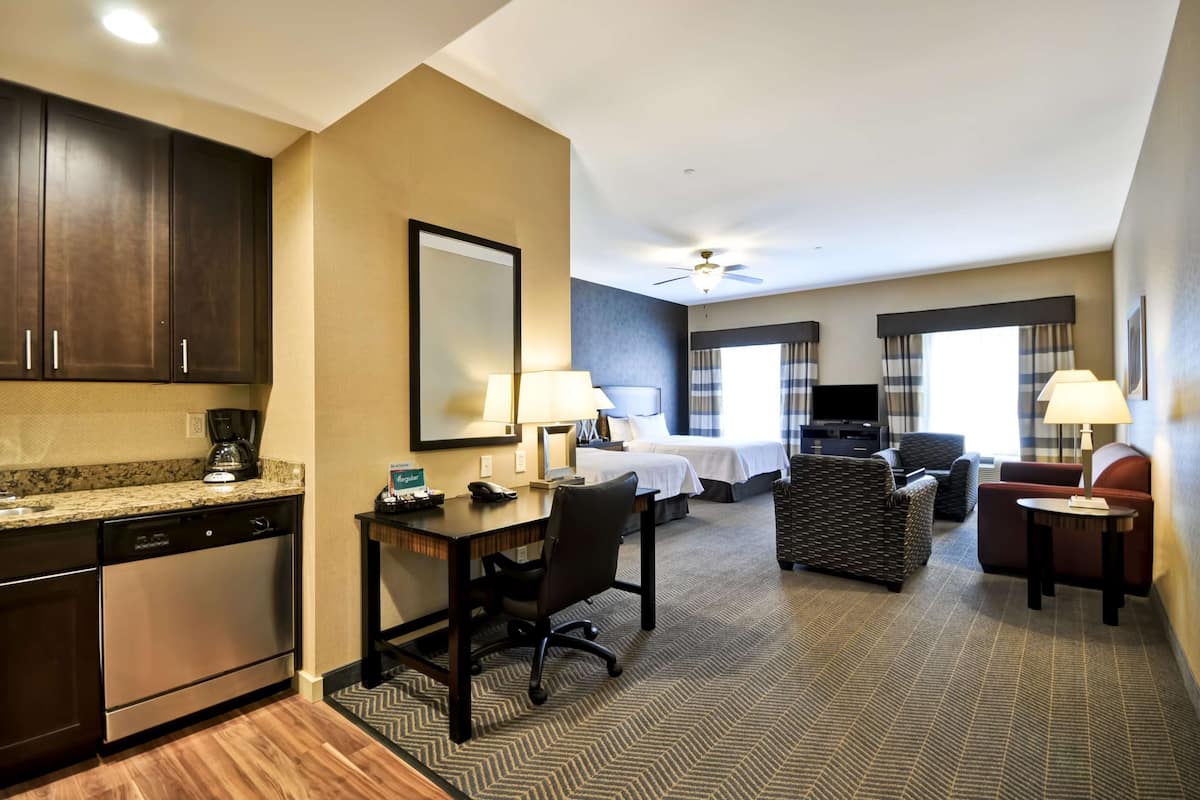 Une chambre d’hôtel équipée d’une kitchenette, d’un lit, d’un bureau avec une chaise, d’une télévision et d’un coin salon avec un canapé et des fauteuils.