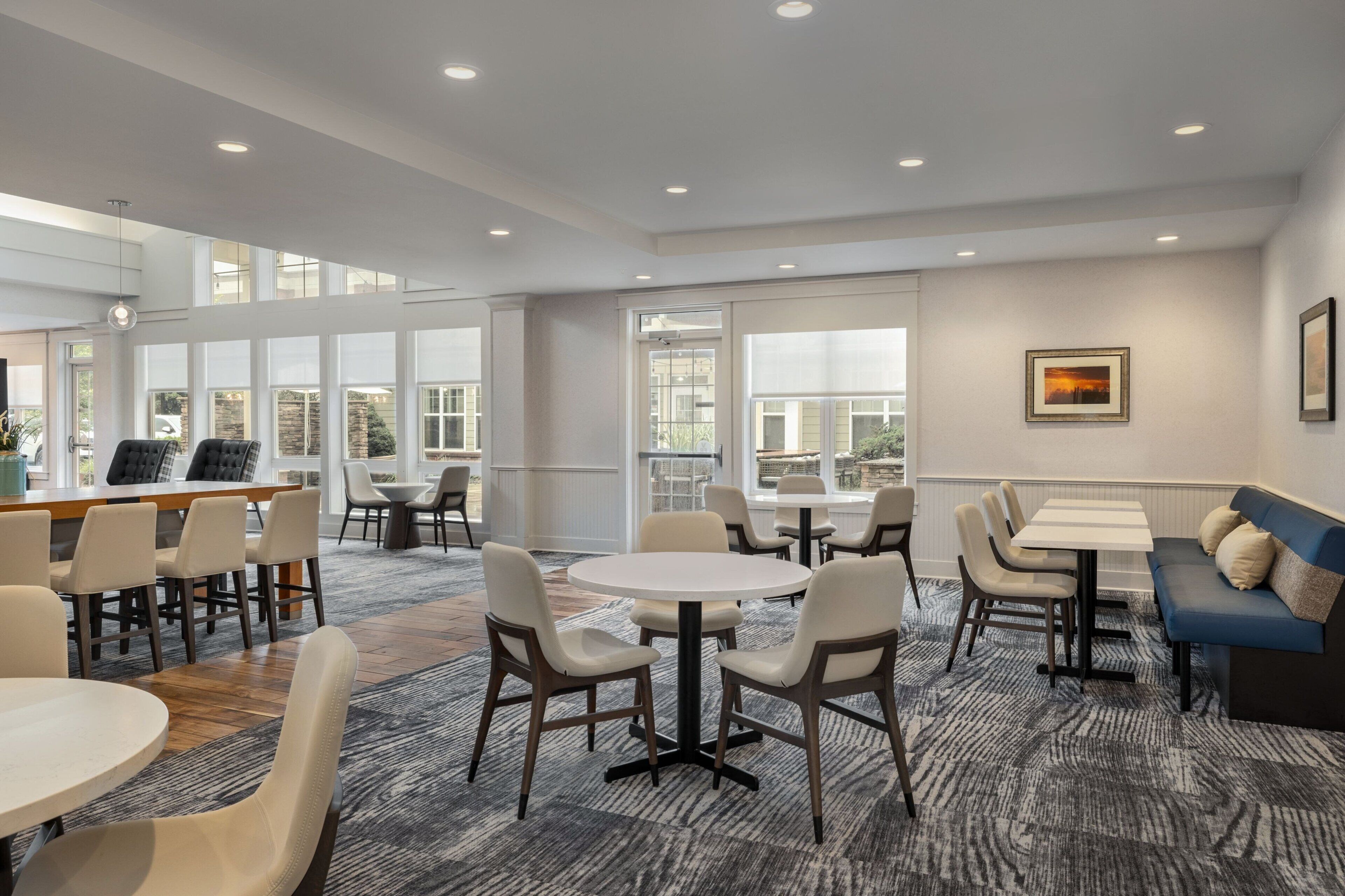 A spacious hotel lobby with multiple seating areas, including round tables and a sofa, and large windows providing natural light.