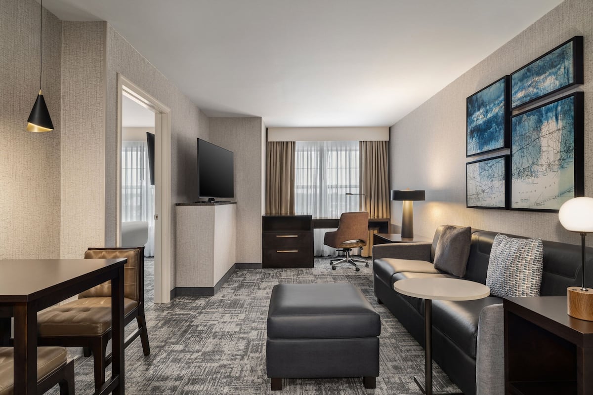 A hotel room with a dark leather sofa, a small round table, a dark ottoman, a TV, a desk with drawers, a chair, and framed artwork on the wall.