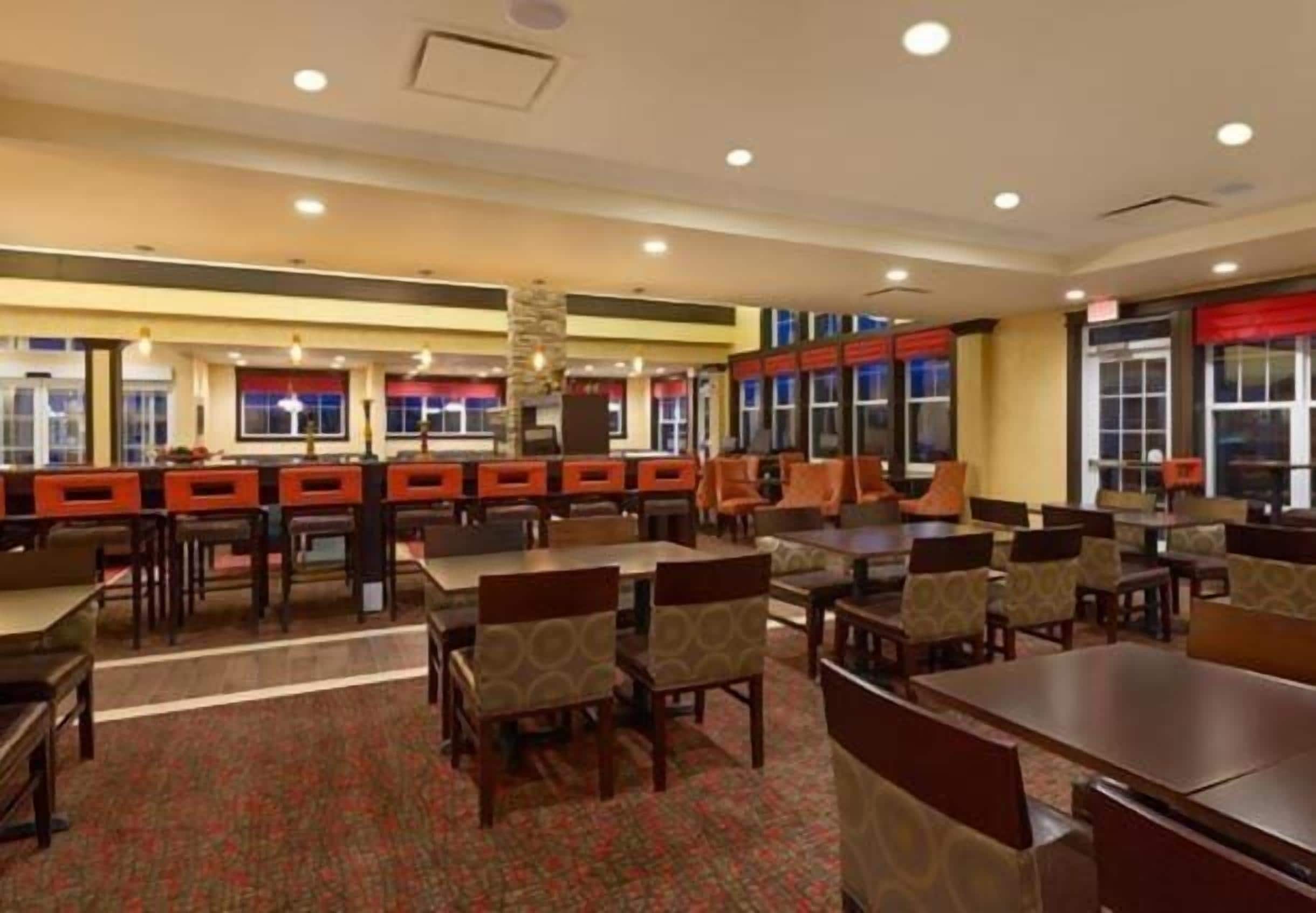 A hotel lobby with multiple dining tables and chairs, a patterned carpet, and large windows with red curtains.