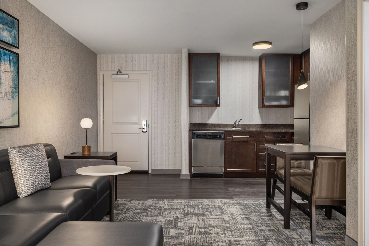 Un espacio habitable compacto con sofá, una pequeña mesa redonda, una zona de cocina con lavavajillas y refrigerador, y una mesa de comedor con sillas.