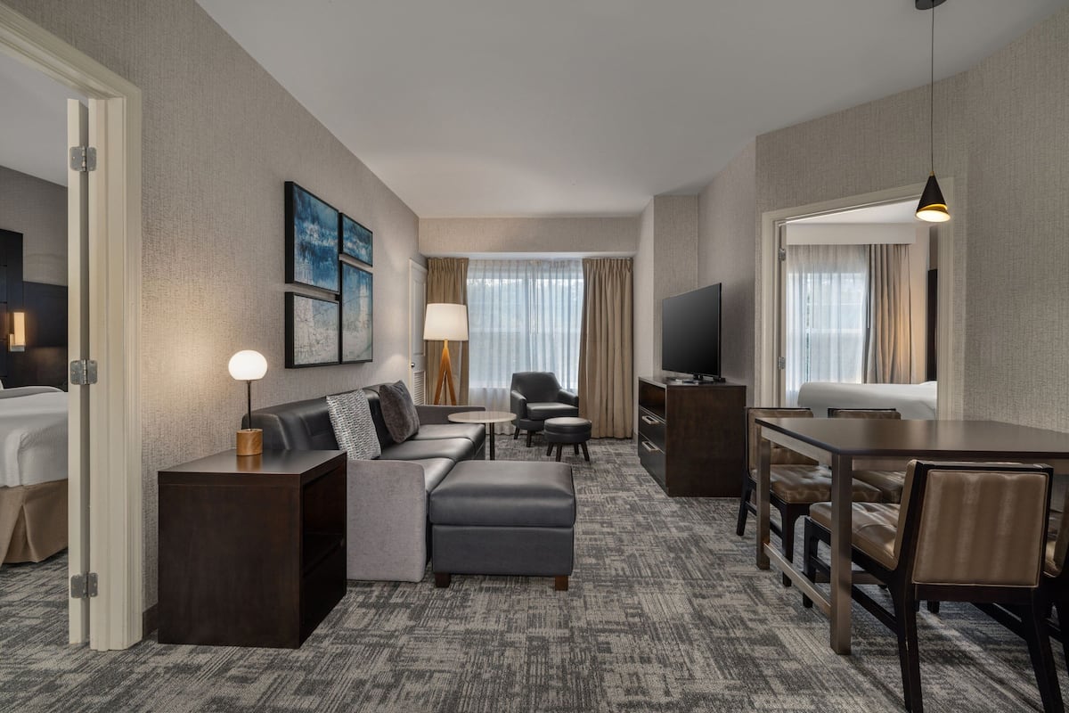 A hotel room with a sofa, ottoman, dining table, chairs, a TV, a lamp, a side table, and a window with curtains.