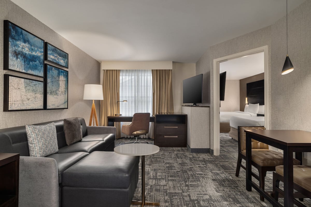 A modern hotel room with a sofa, a small round table, a desk, a TV, and a bed.