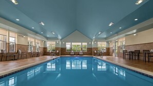 Una amplia piscina interior con agua cristalina, rodeada de sillones y mesas, con un techo alto y ventanas grandes.