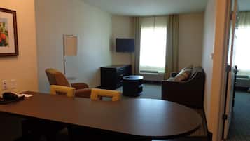 A hotel room with a dining table, two armchairs, a sofa, a flat-screen TV, and a large window with curtains.
