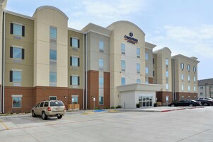 Un hotel de varios pisos con fachada plana, entrada central y un estacionamiento con algunos vehículos.