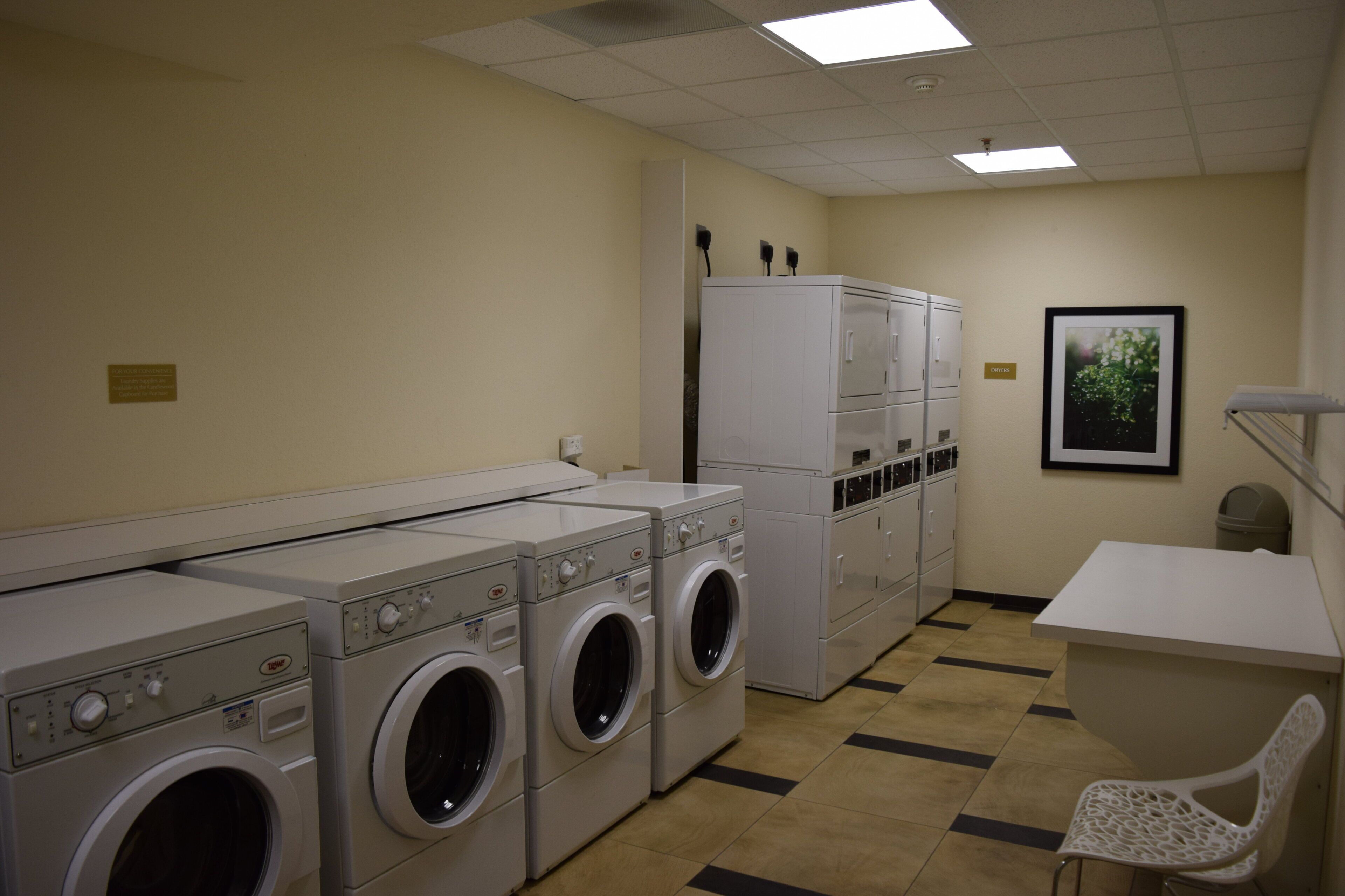 A laundry room with multiple washing machines and dryers, a white chair, and a framed picture on the wall.