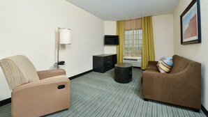 Habitación de hotel con un sofá color beige, un sofá marrón, una mesita de noche negra, una lámpara de pie, un televisor en la pared y una ventana con cortinas amarillas.
