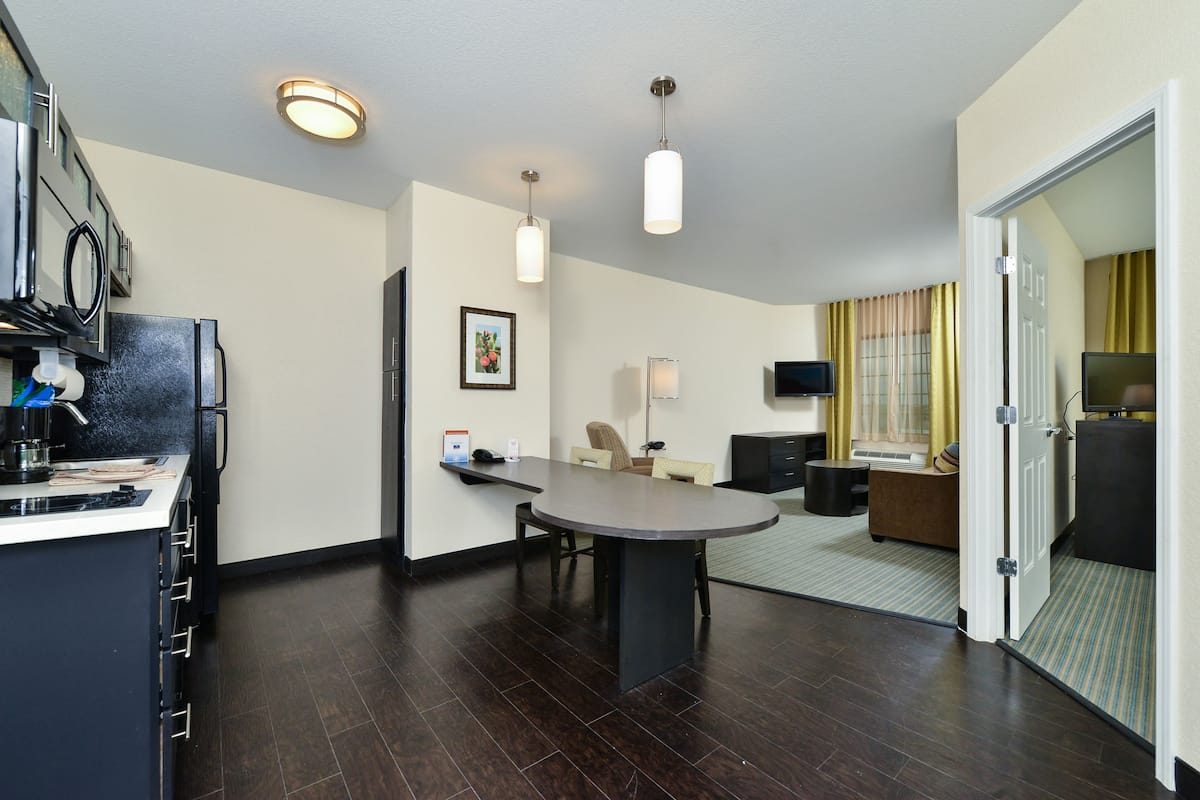 A modern hotel room with a kitchen area, a dining table, a sofa, and a TV.