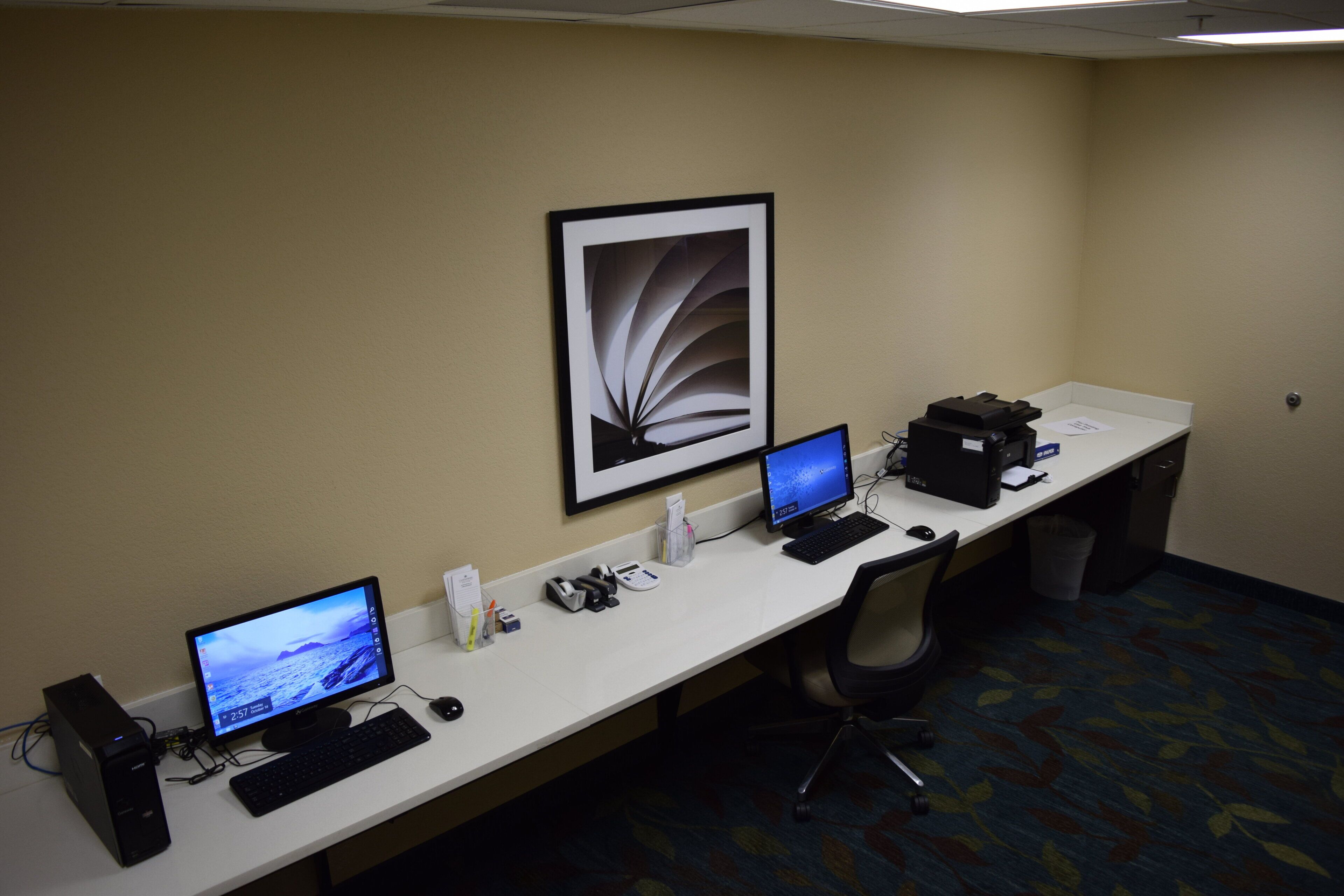 A hotel workspace with three computer monitors, a printer, and a desk lamp.
