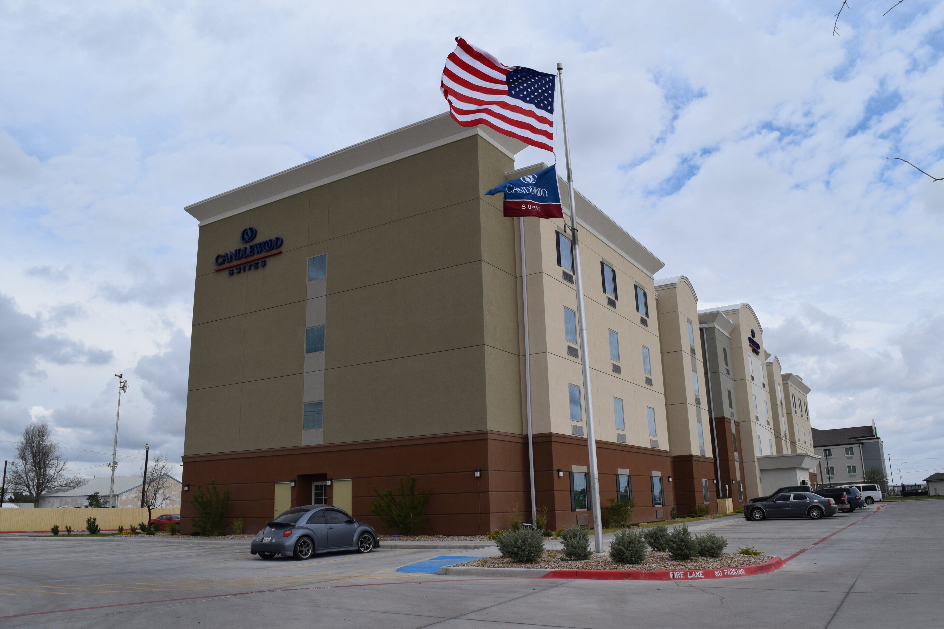 A hotel with a flagpole, a sign for 'Candlewood Suites,' and a parking lot with cars.