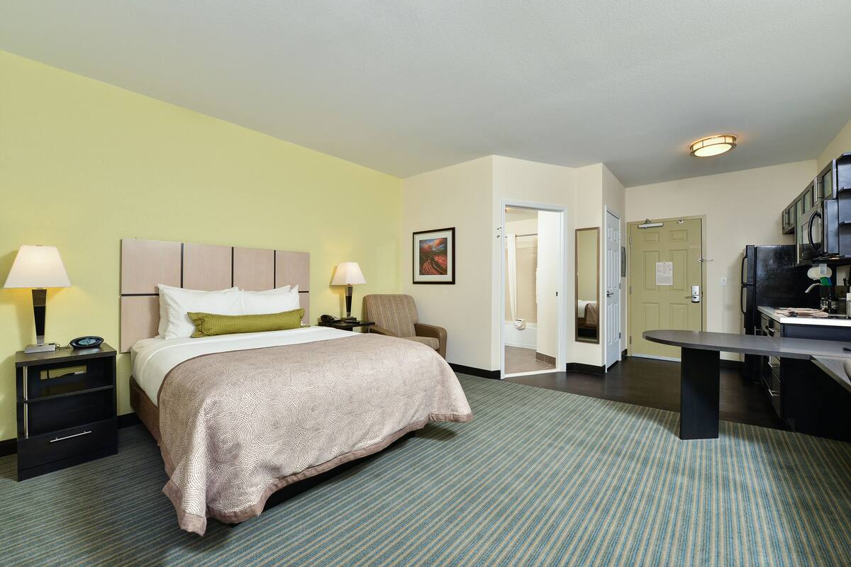 A hotel room with a bed, bedside tables, a chair, a desk, and a kitchenette.