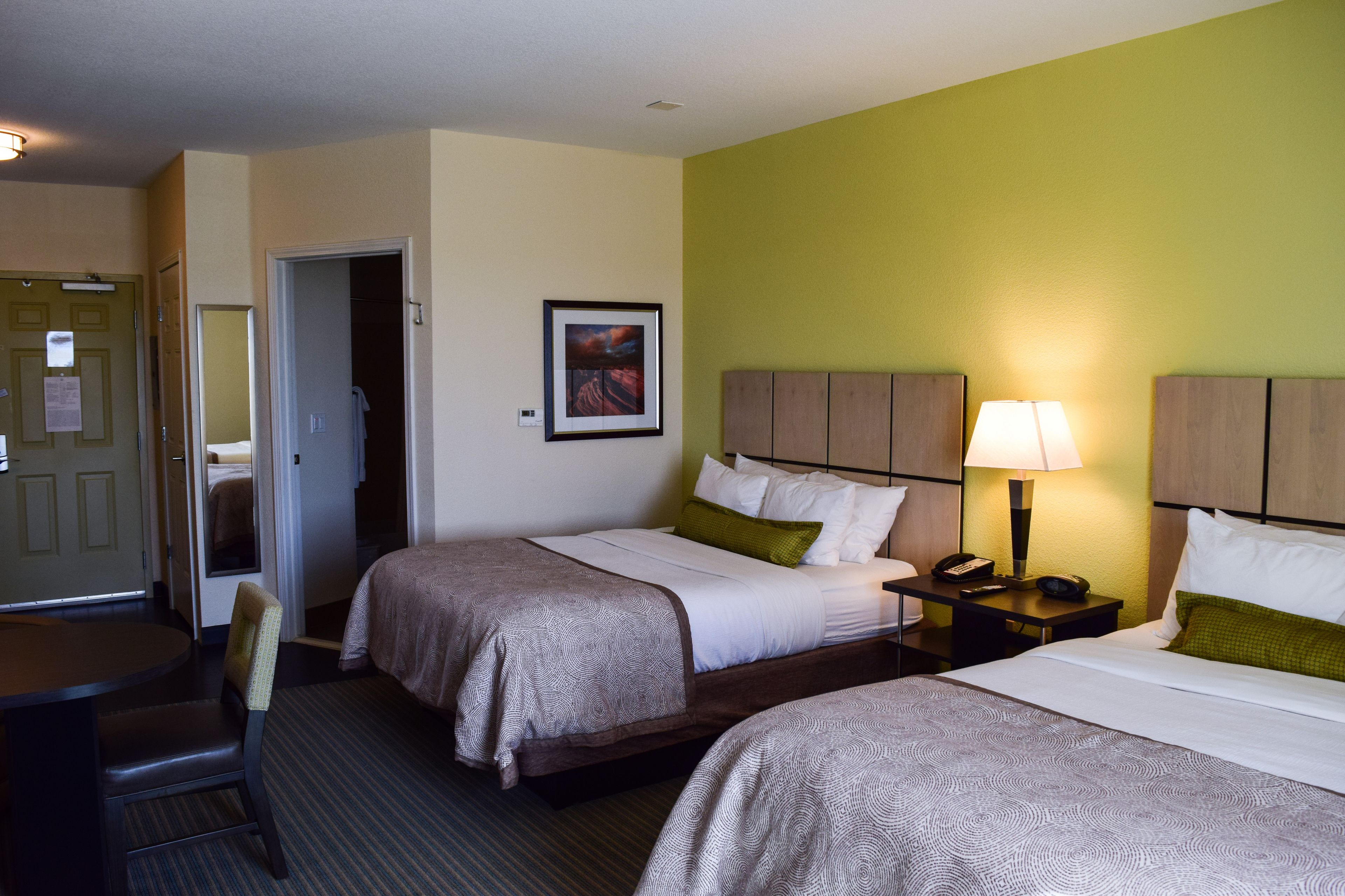 A hotel room with two beds, a desk, and a chair. There is a door leading to another room and a framed picture on the wall.
