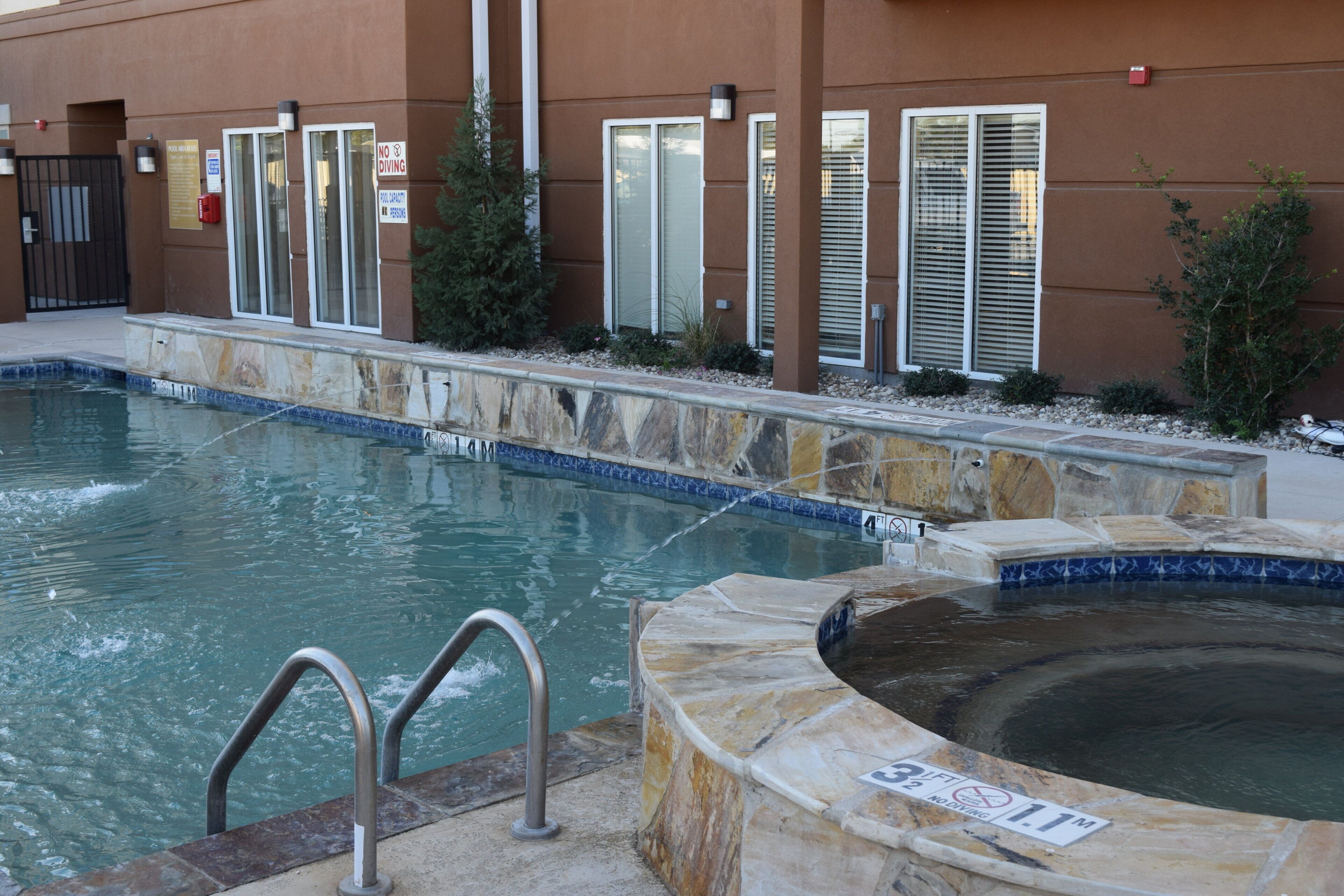 A swimming pool with a hot tub, surrounded by a stone deck and a building with windows and a gate.