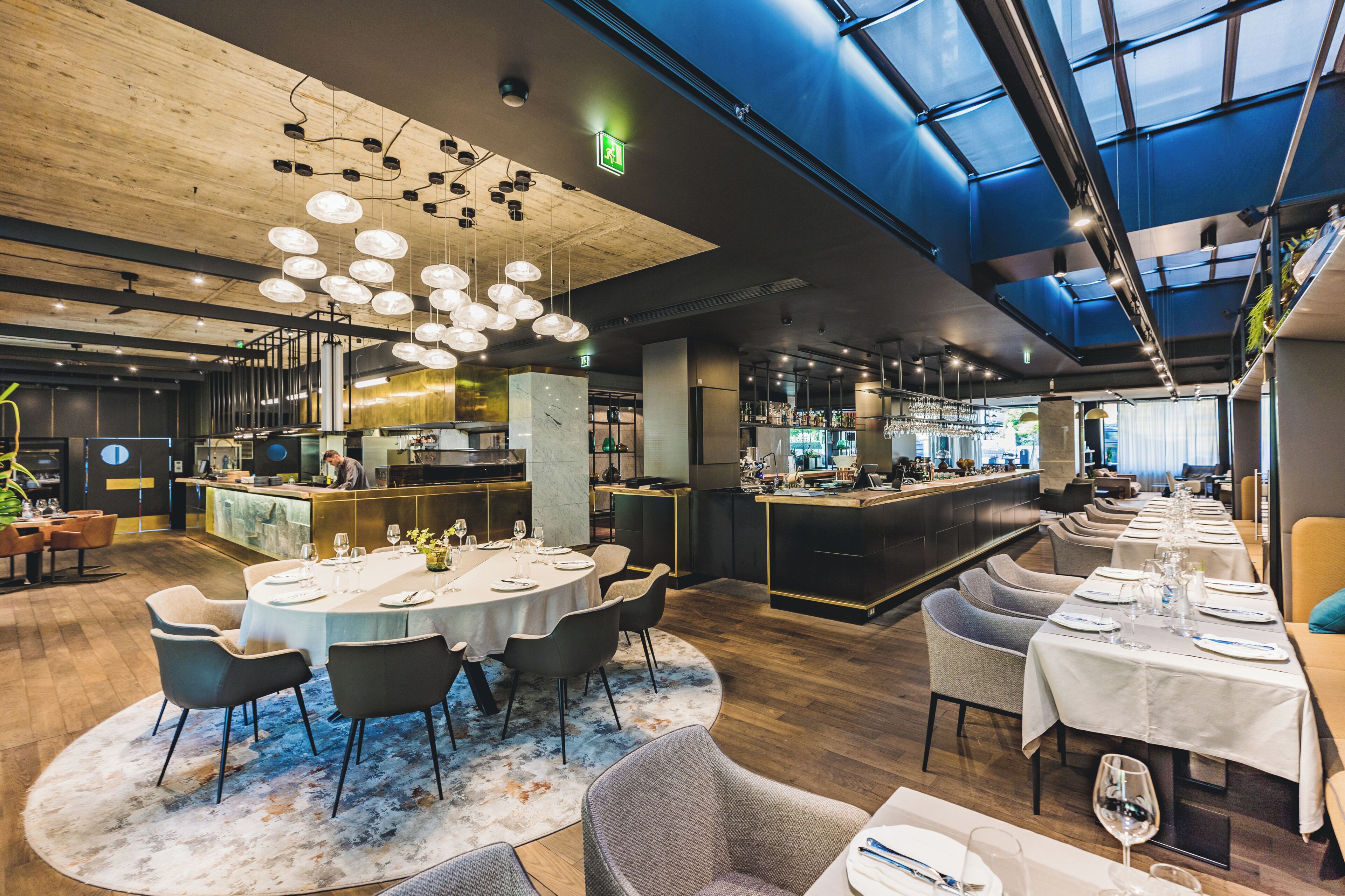 A modern restaurant with round tables, a bar area, and a high ceiling with exposed beams.