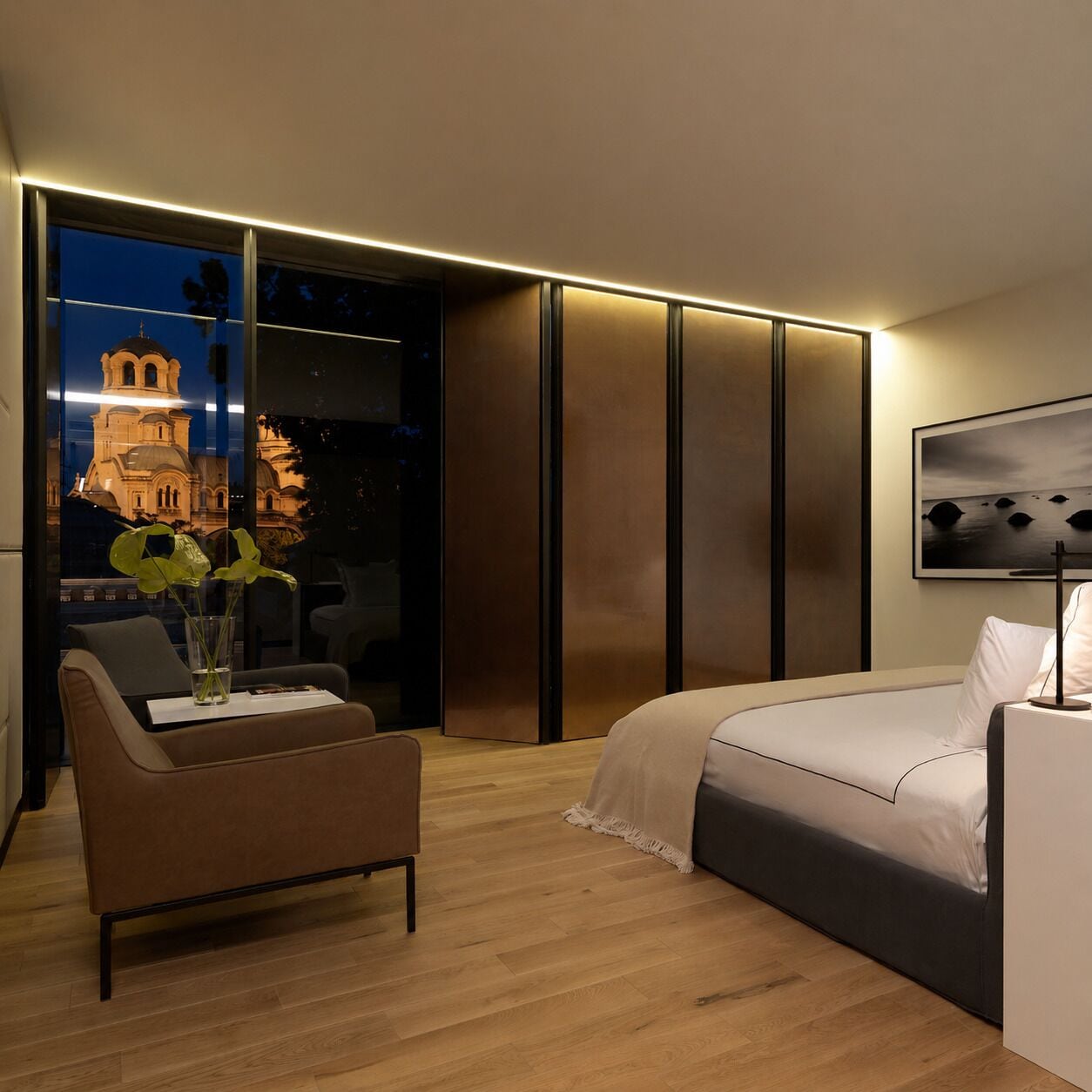 A modern hotel room with a bed, a chair, a vase with flowers, a window with a cityscape view, and a framed picture on the wall.